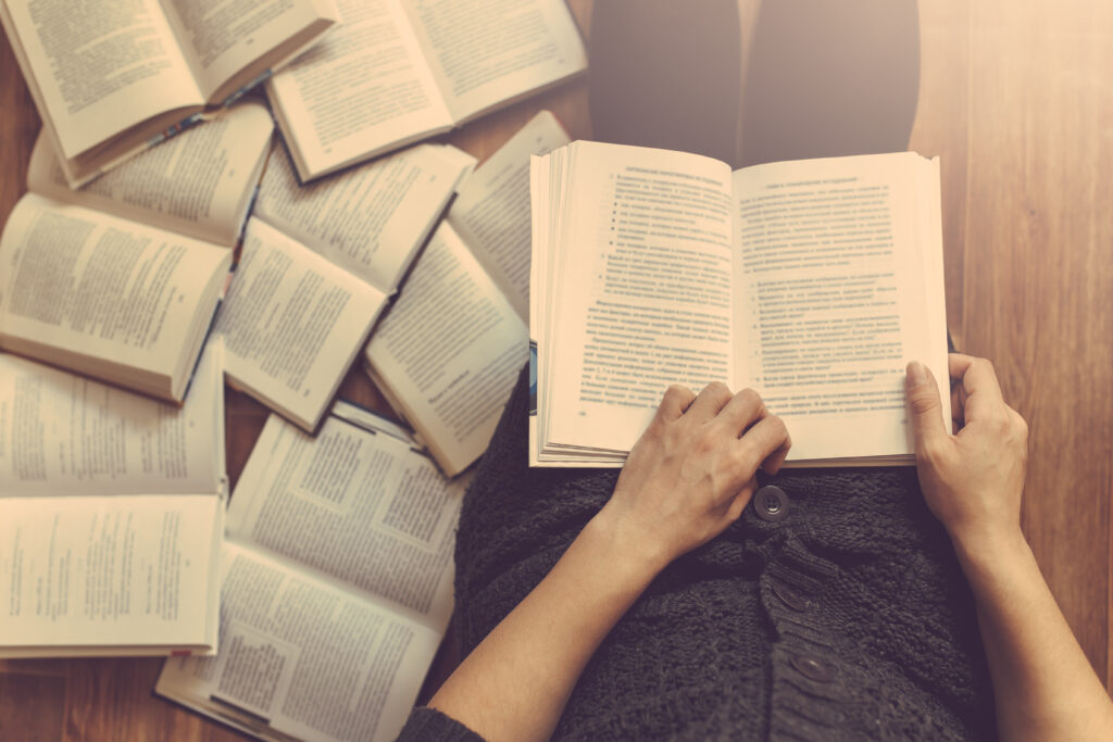 A person's hands holds an open book next to a pile of opened books scattered on a hardwood floor.