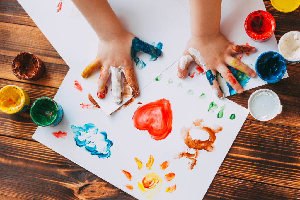 A child uses their hands to finger paint with yellow, red, green, blue, and white paint on a few sheets of white paper. 