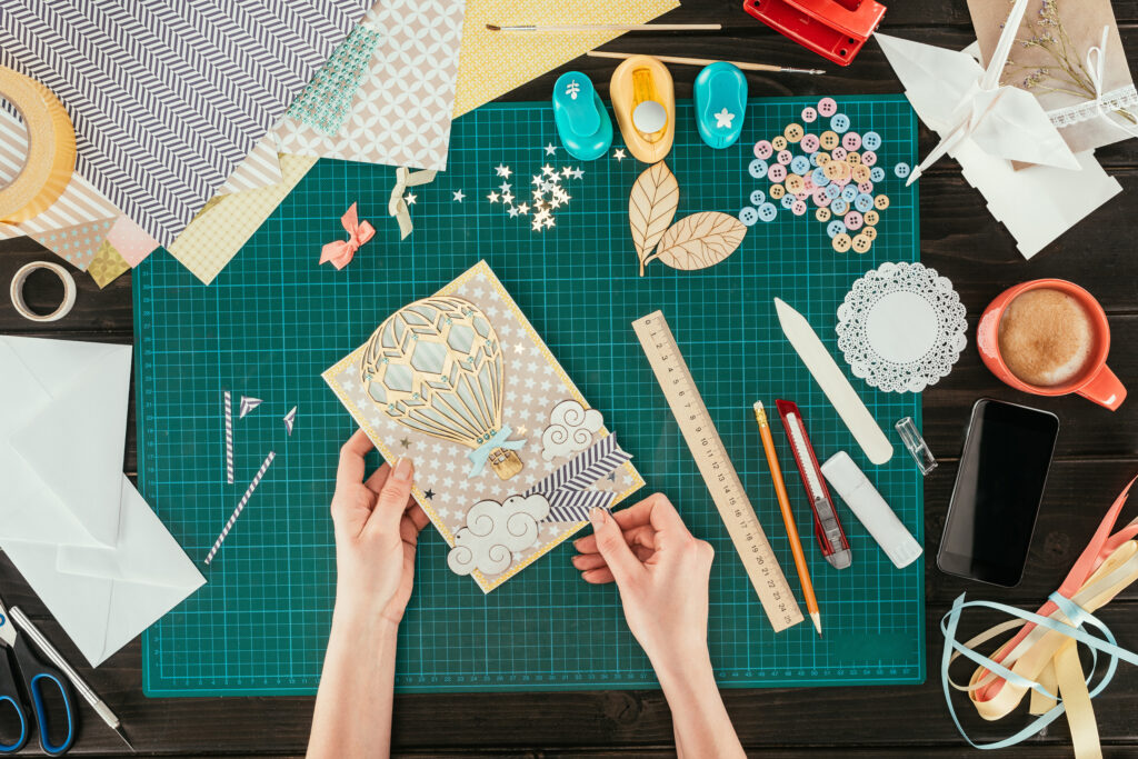 A pair of hands is scrapbooking with buttons, paper, ribbon, and other art supplies to make a colorful greeting card with the image of a hot air balloon on the card's cover. 
