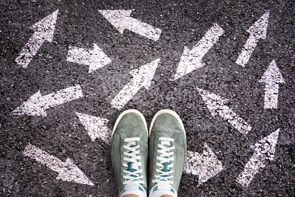 A pair of grey sneakers on black asphalt with white painted arrows pointing in various directions.