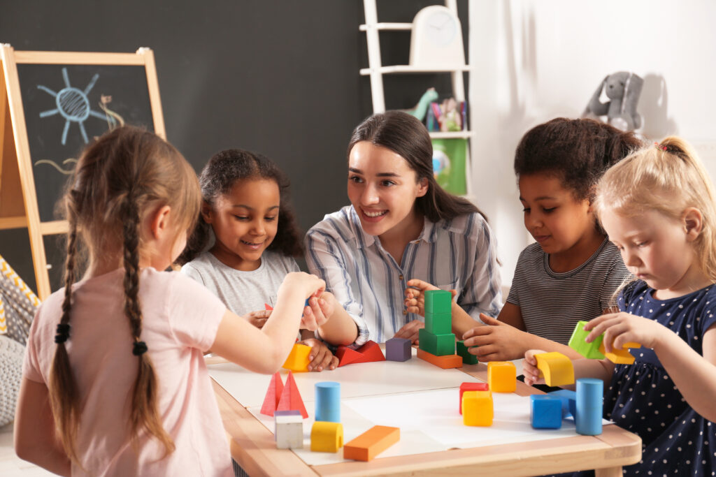 A smiling female teacher seated at a table with four female students playing with blocks.