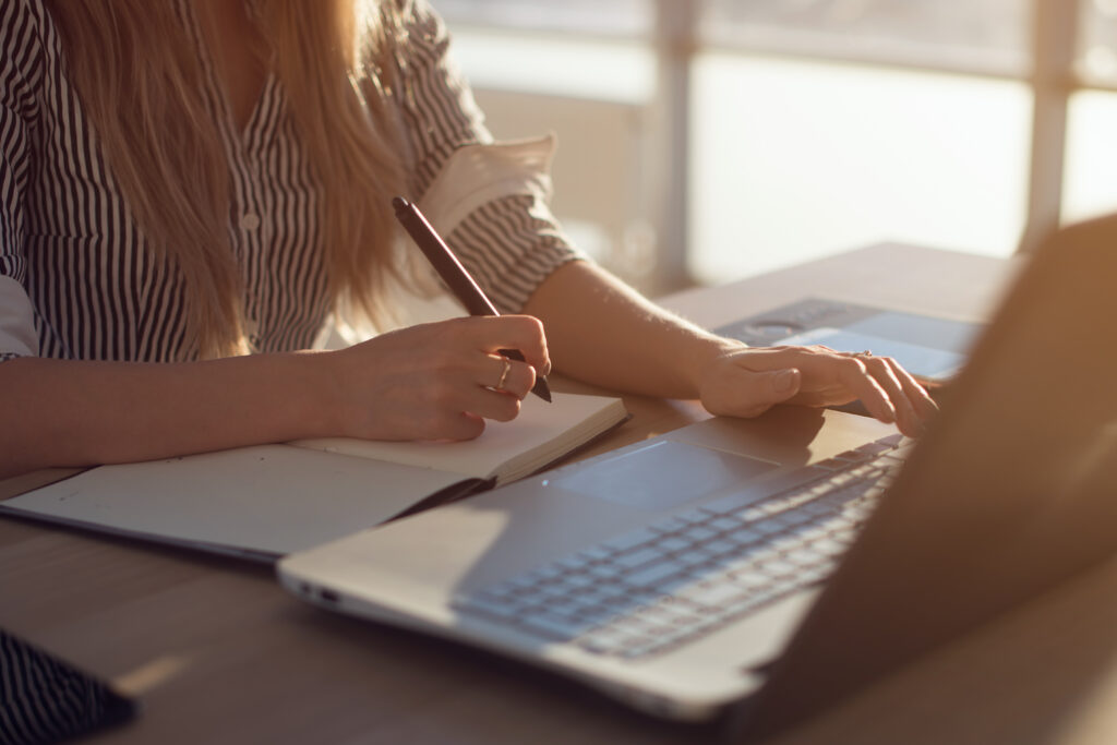 A photograph of a woman writing in a journal while using a laptop in front of her. 